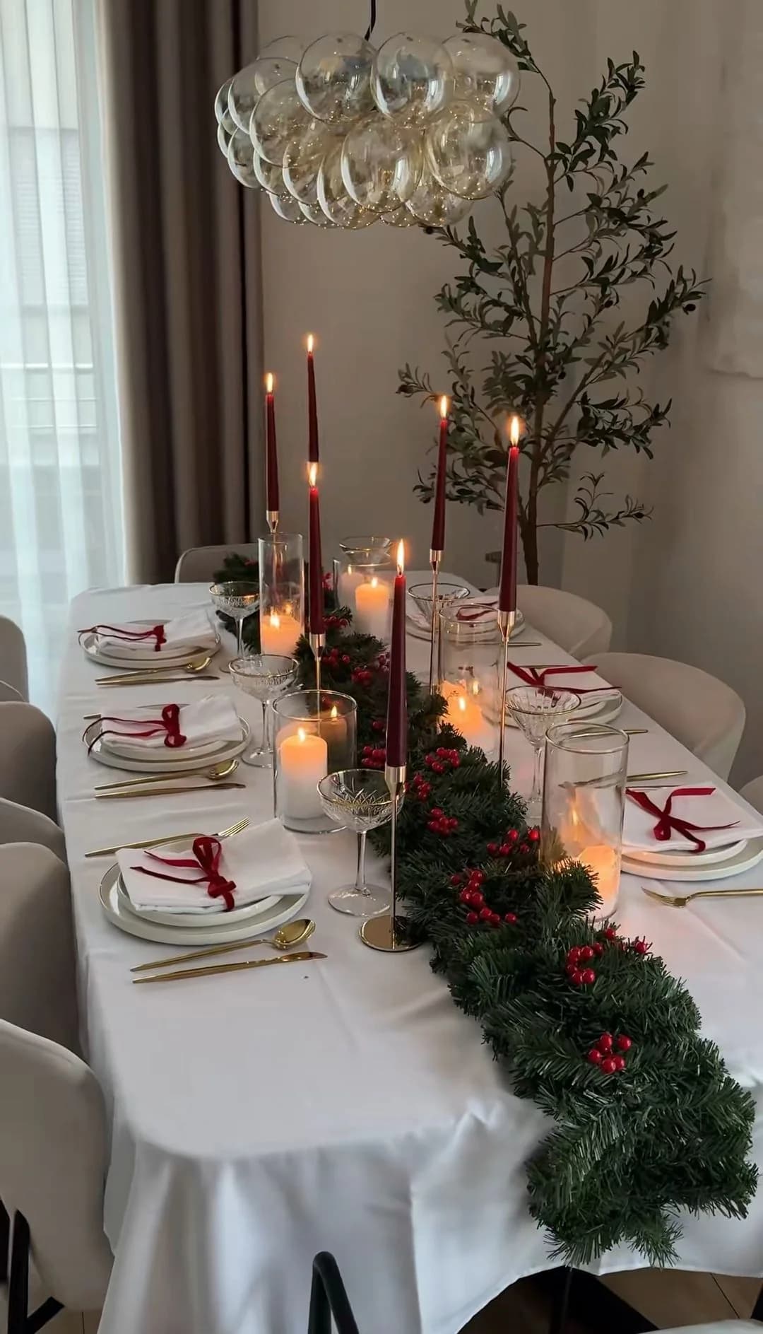 Mesa de comedor con mantel blanco, una guirnalda verde con frutos rojos al centro, velas largas color vino, velas en vasos de cristal y servilletas blancas atadas con listón vino