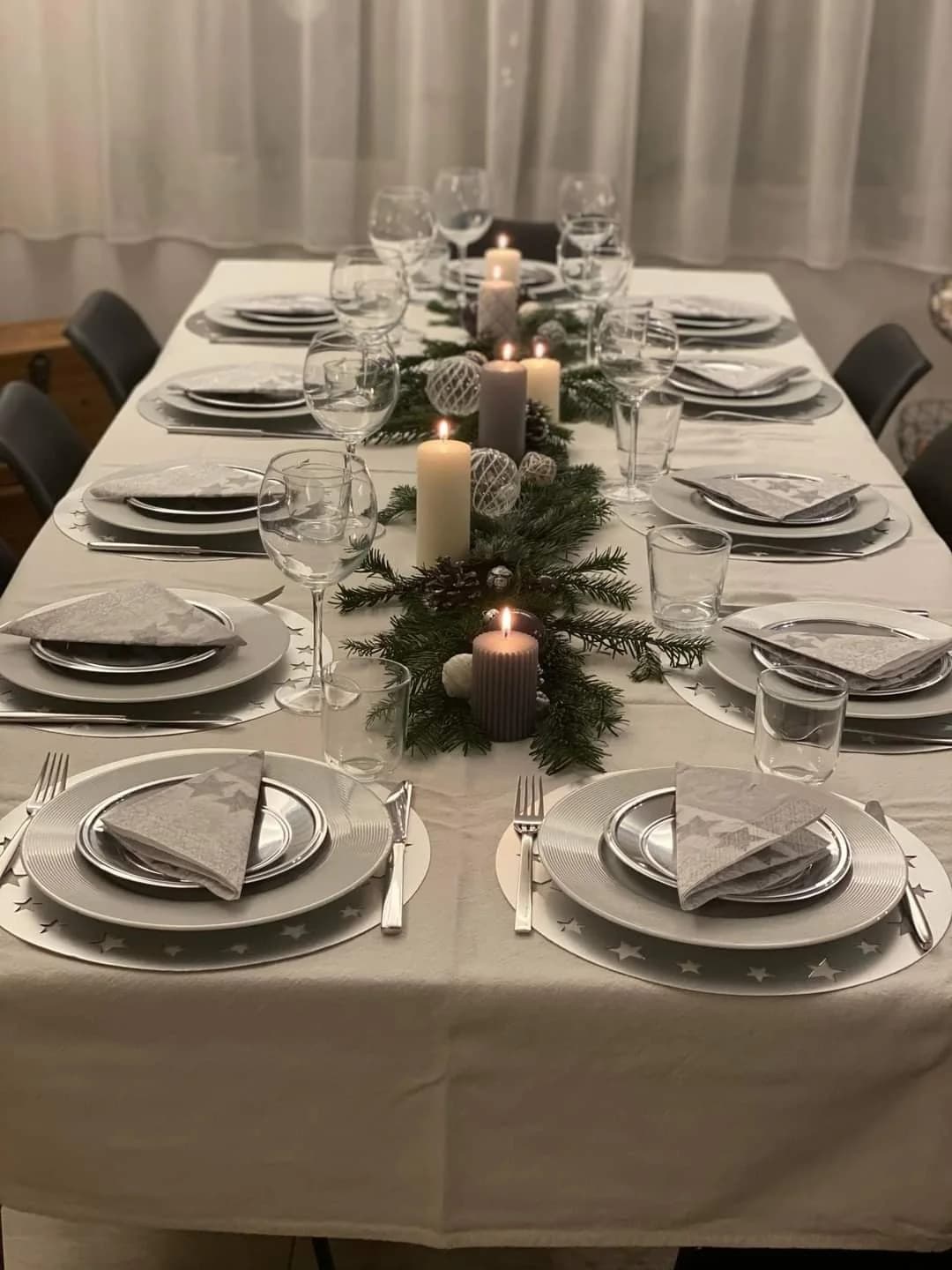 Mesa de comedor con mantel gris claro, platos base plateados, servilletas grises y un centro de ramas de pino con velas gruesas en gris y blanco y esferas de cristal