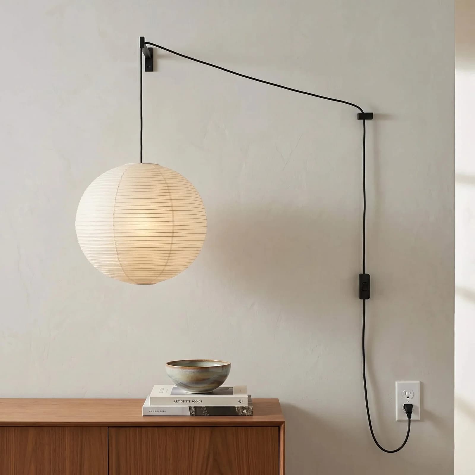 Lámpara de esfera de papel sostenida por un brazo de metal negro en la pared, con el cable hasta un contacto