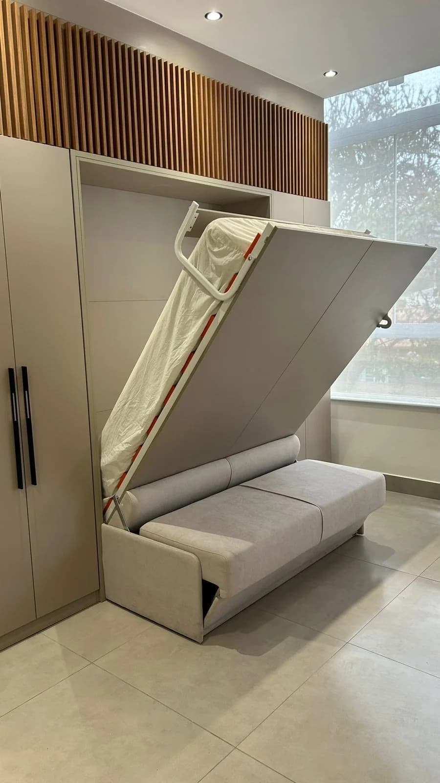 Cama abatible a medio bajar desde un mueble de pared gris, con un sofá integrado al frente, junto a un clóset alto
