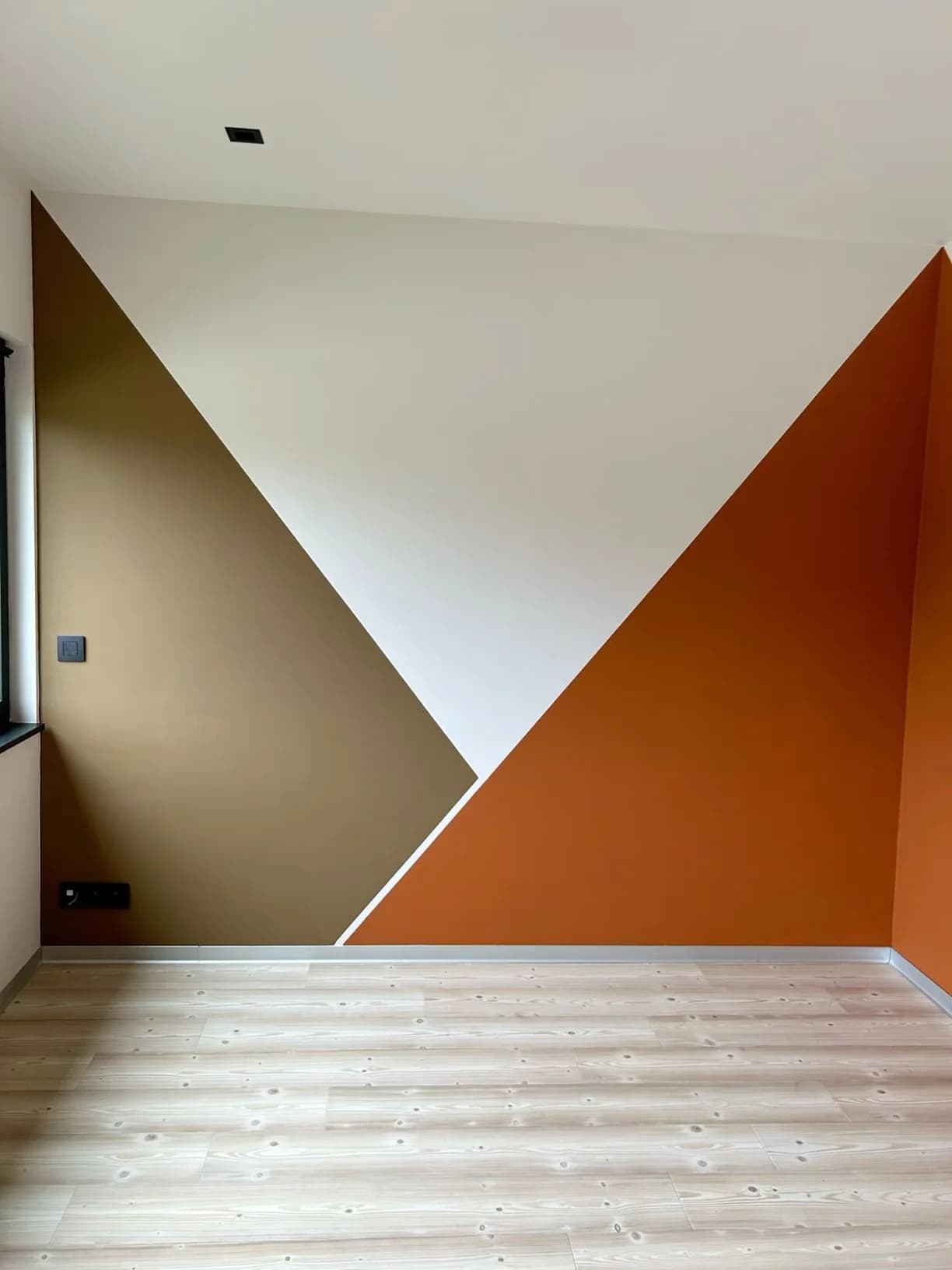 Esquina de un cuarto vacío con triángulos grandes pintados en café claro, blanco y naranja quemado