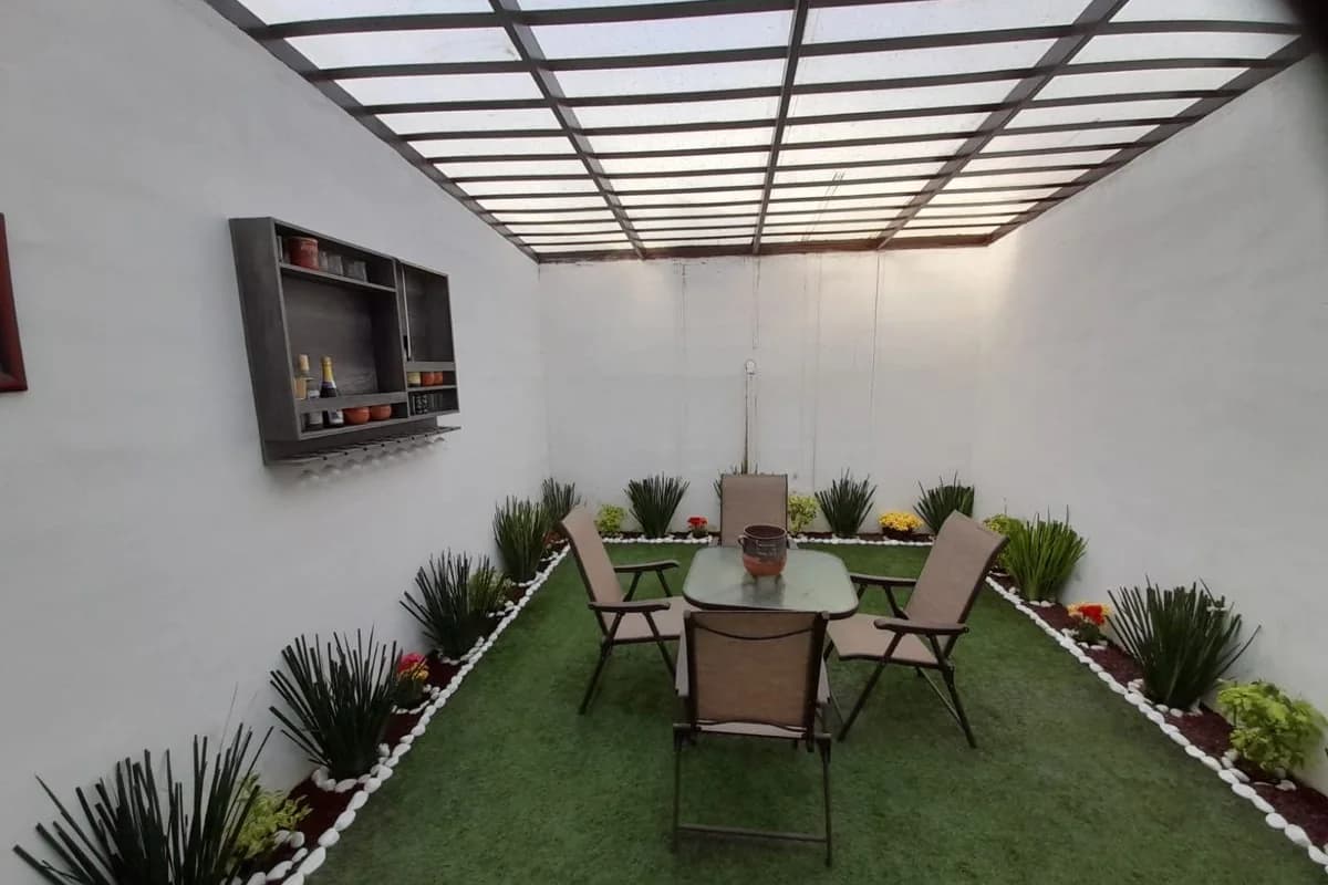 Patio pequeño con muros blancos, techo de policarbonato, pasto sintético, jardinera perimetral con plantas y mesa con cuatro sillas