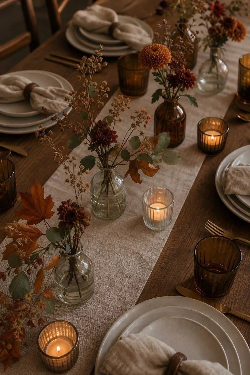 Linen runner on a wooden table with small glass vases of chrysanthemums and dried stems, amber glasses and tea lights between the plates