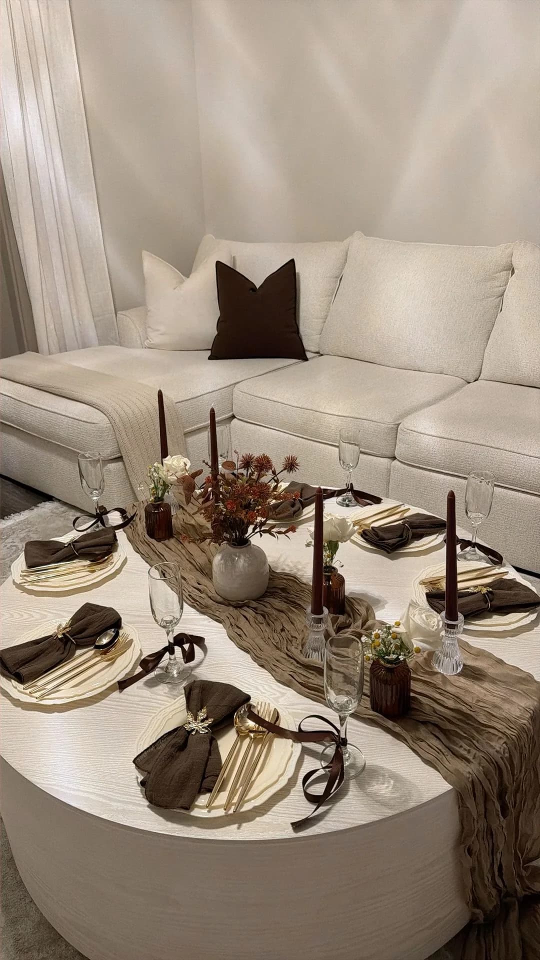 Round white coffee table in front of a sofa set for four with a gauze runner, brown napkins tied with ribbon, a small vase and dark taper candles