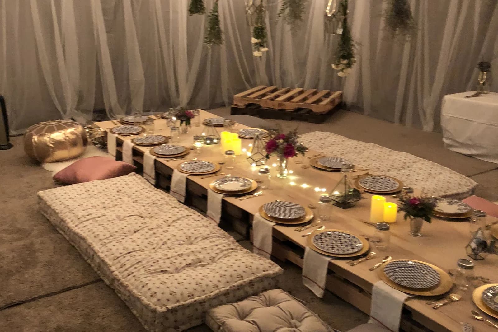 Long low table made from wooden pallets set for dinner with floor cushions, candles and a strand of warm lights down the middle