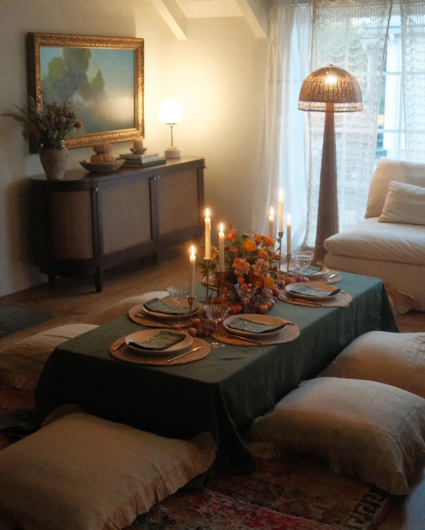Low table covered with a dark green cloth and set for dinner with taper candles and fall flowers, surrounded by large floor cushions on a rug
