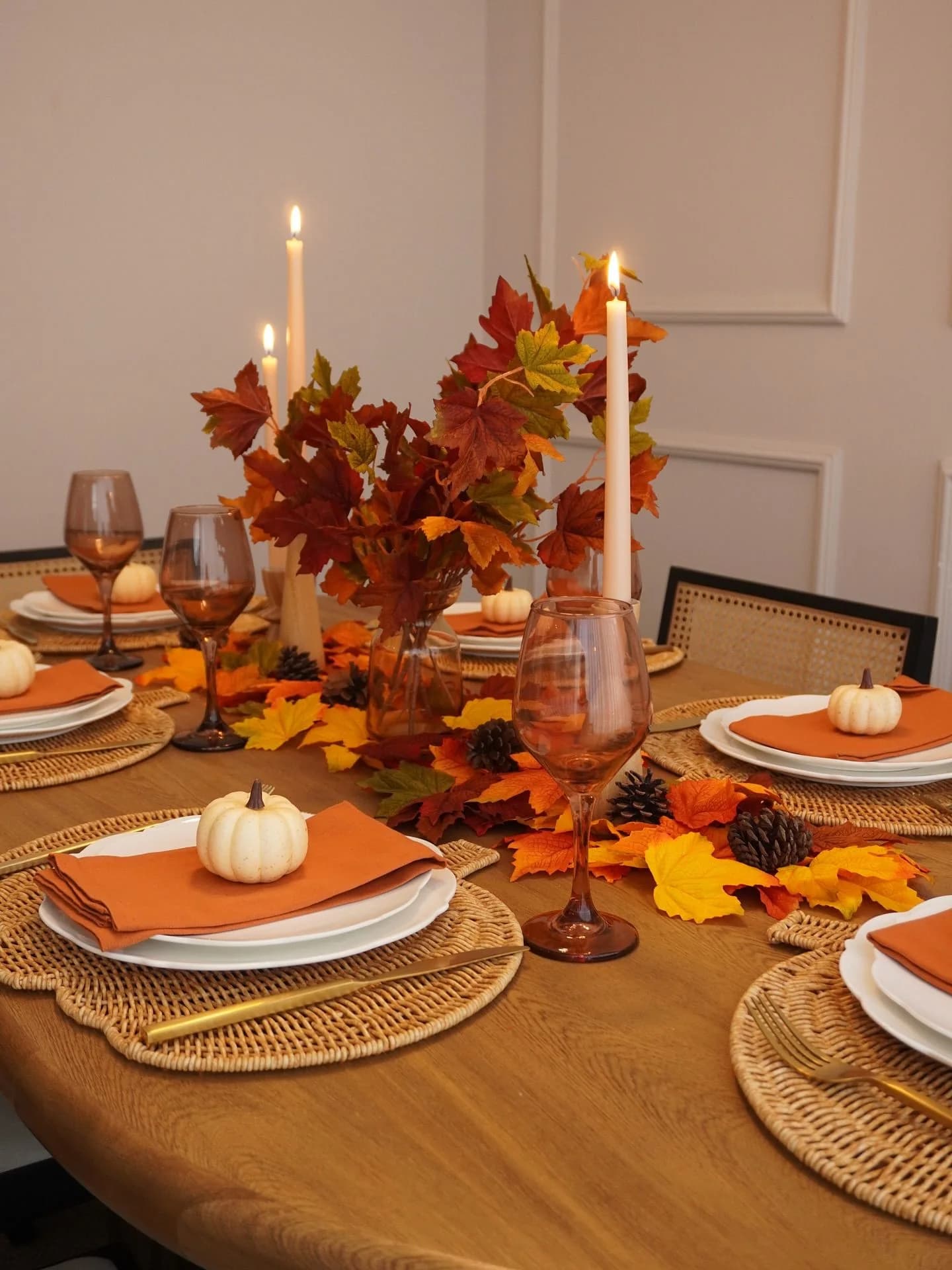Round wooden table with maple leaves, pine cones, taper candles, amber glasses and a white mini pumpkin on each orange napkin