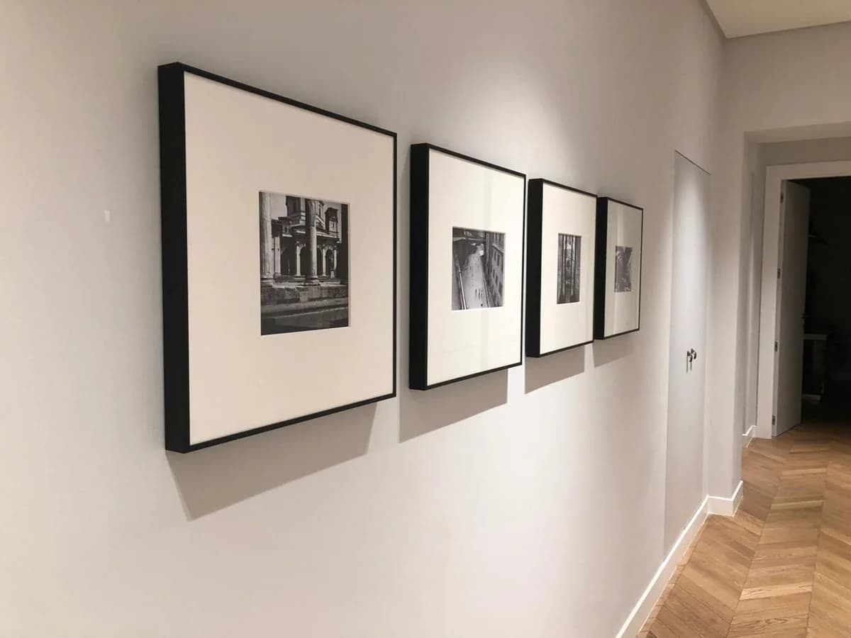 Hallway wall with a row of four black frames with wide white mats holding small black-and-white architecture photographs, above a herringbone wood floor