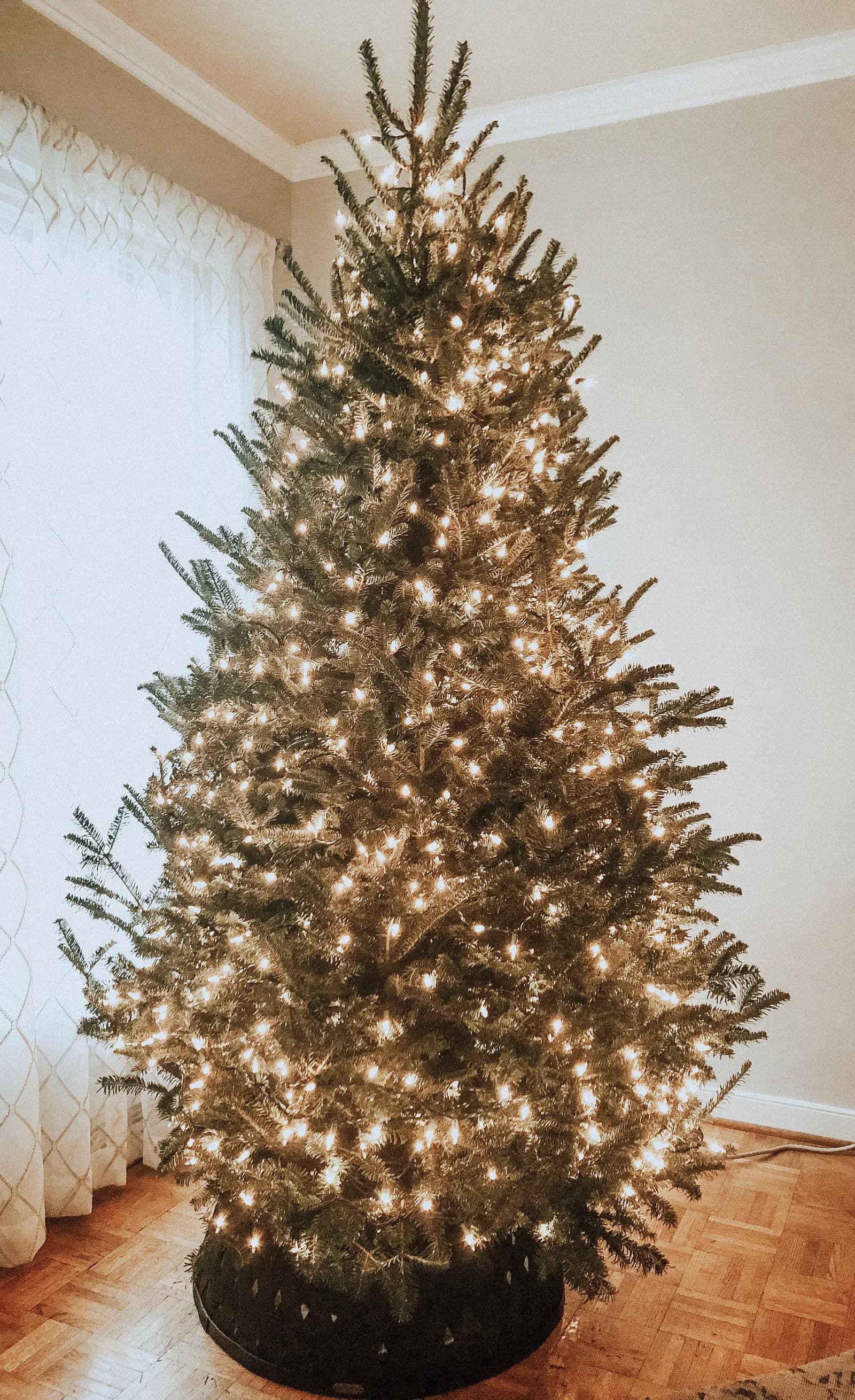Real fir tree with warm white mini lights and no ornaments in a room with parquet floor and sheer white curtains