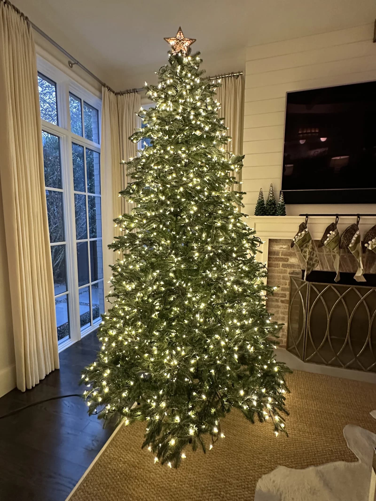 Tall artificial Christmas tree covered in warm white mini lights with a star on top and no ornaments, beside cream curtains and a white fireplace