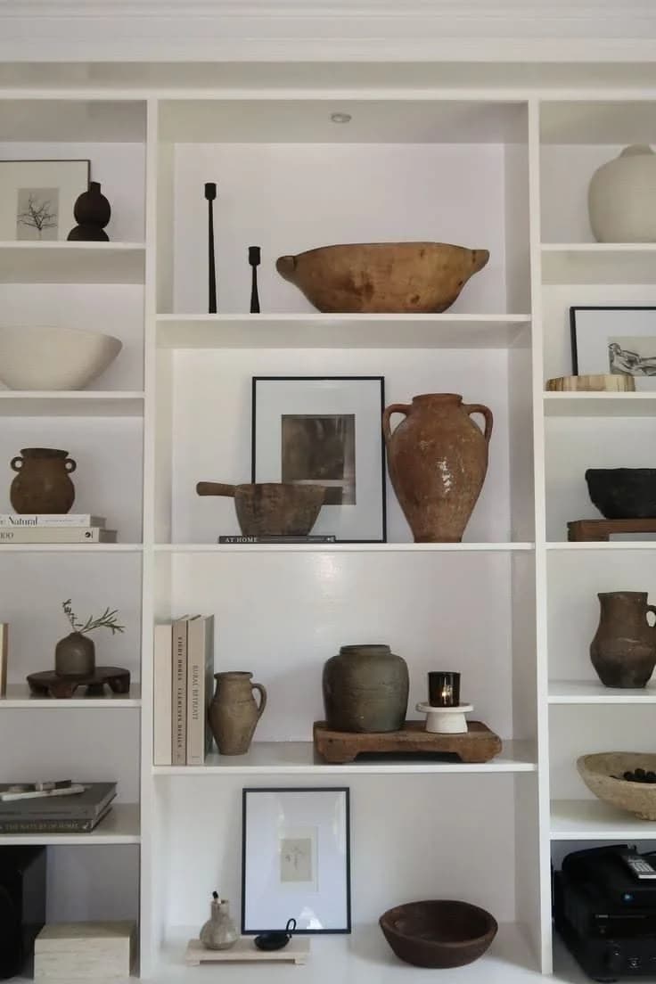 White shelving unit with no books on most shelves, styled with old terracotta jugs, wooden bowls, dark pottery, black candlesticks and two framed drawings