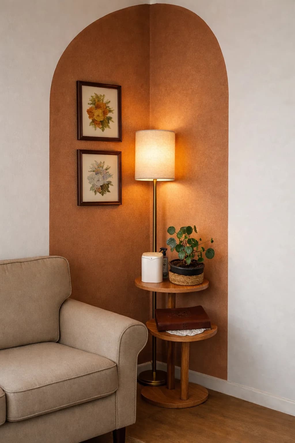 Terracotta arch painted across a corner with two framed botanical prints, a slim floor lamp with a linen shade, a two-tier wooden side table with a plant and a beige armchair