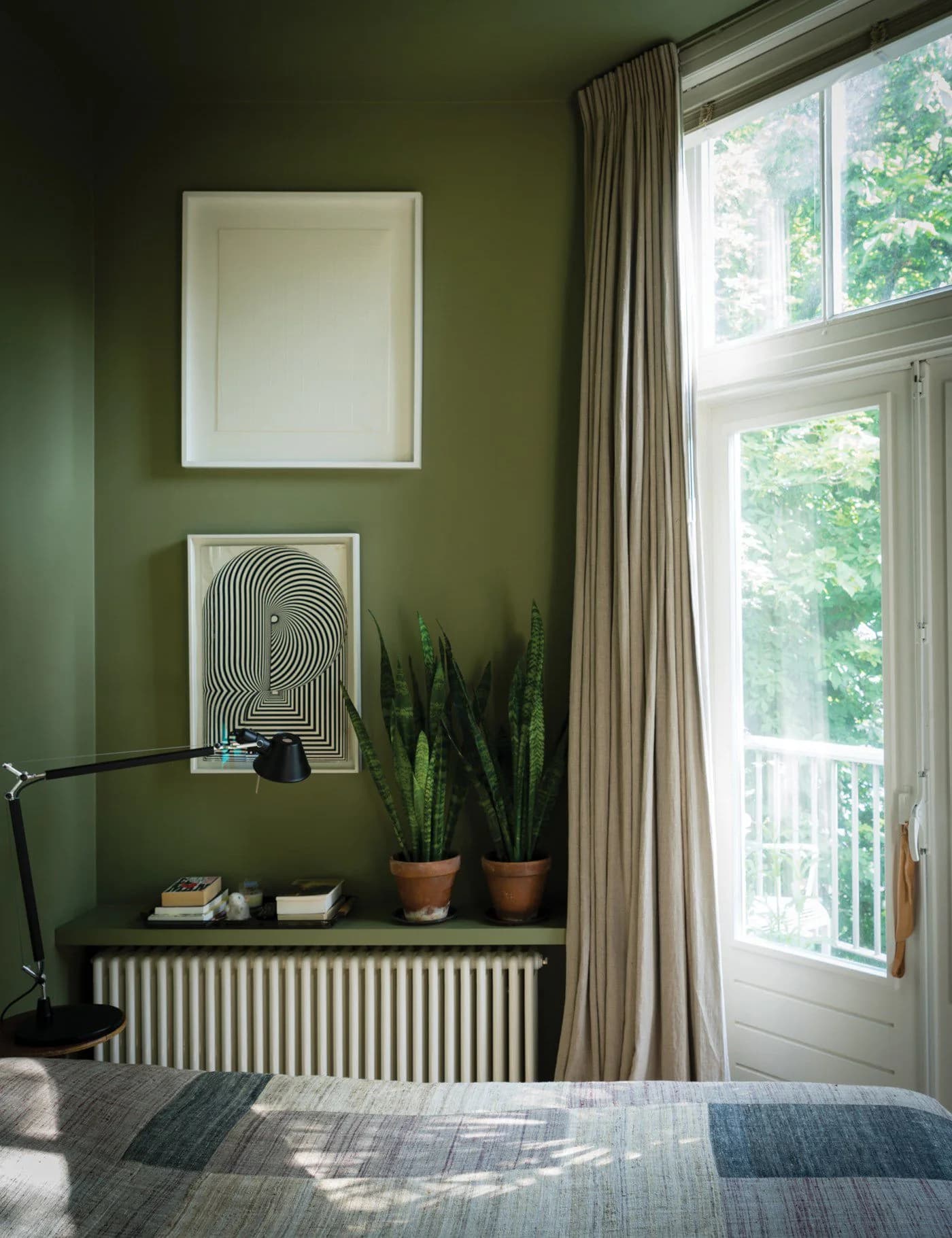 Corner with a moss green wall and ceiling, a long natural linen curtain at a tall window, two prints, snake plants on a shelf above a radiator and a black desk lamp