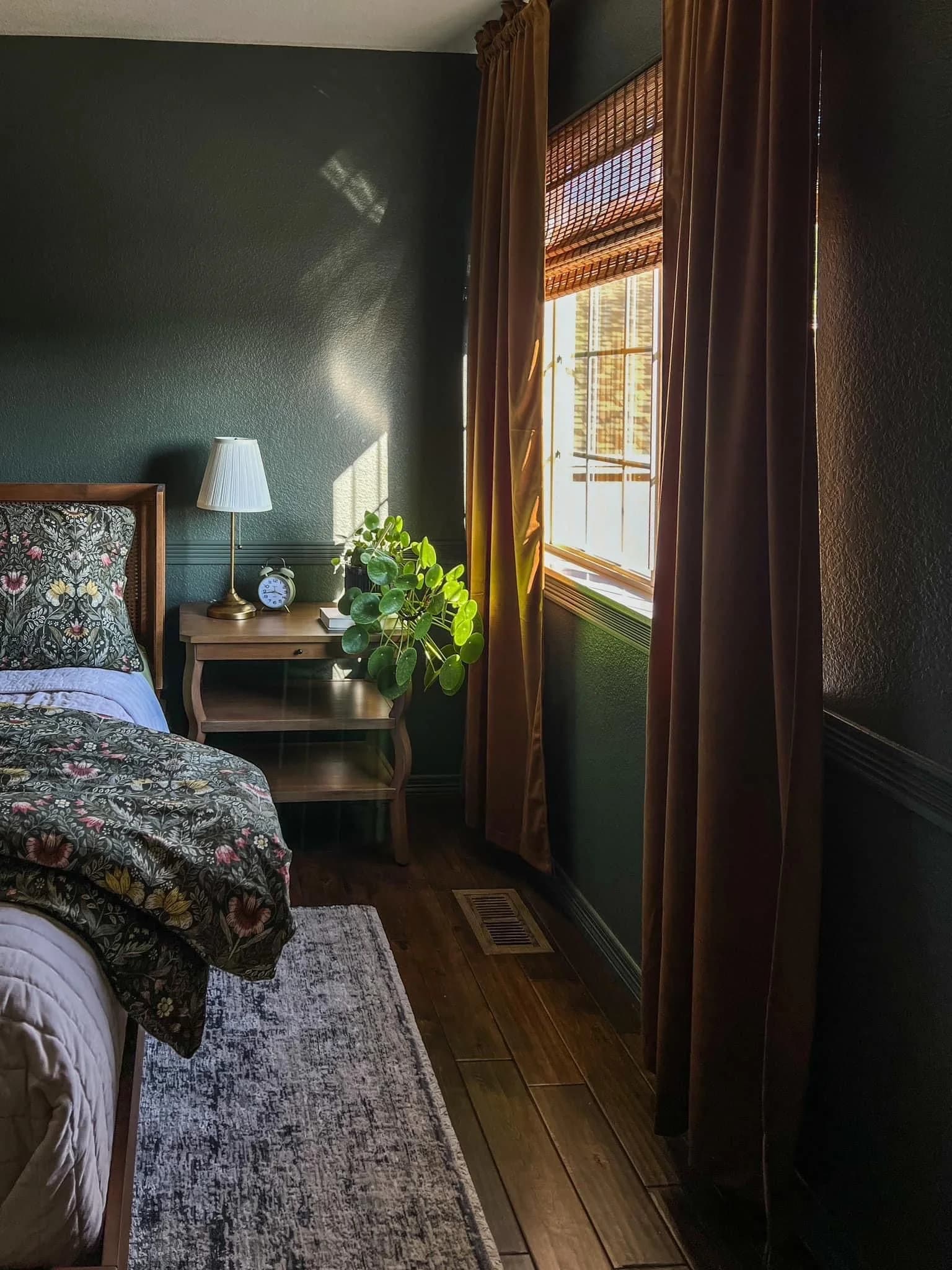 Bedroom with dark green textured walls, long rust brown velvet curtains over a woven wood blind, a wooden bedside table with a pleated lamp and a floral bedspread