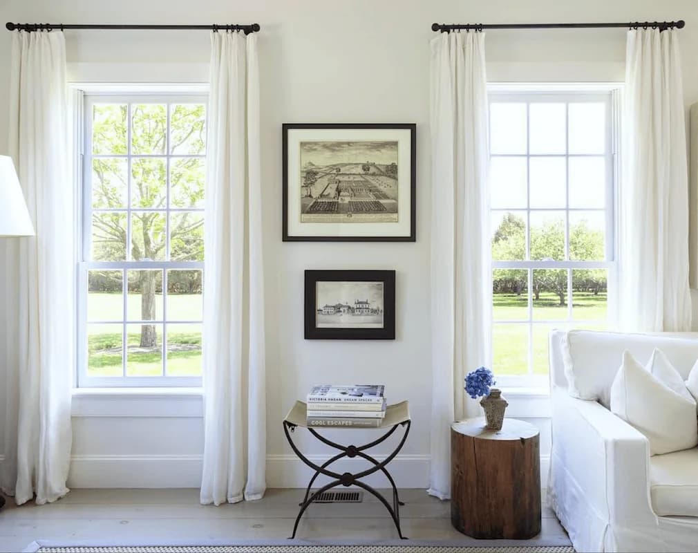 White living room with two sash windows dressed in full-length white curtains on black rods, two framed prints between them, a white sofa and a wooden stump side table