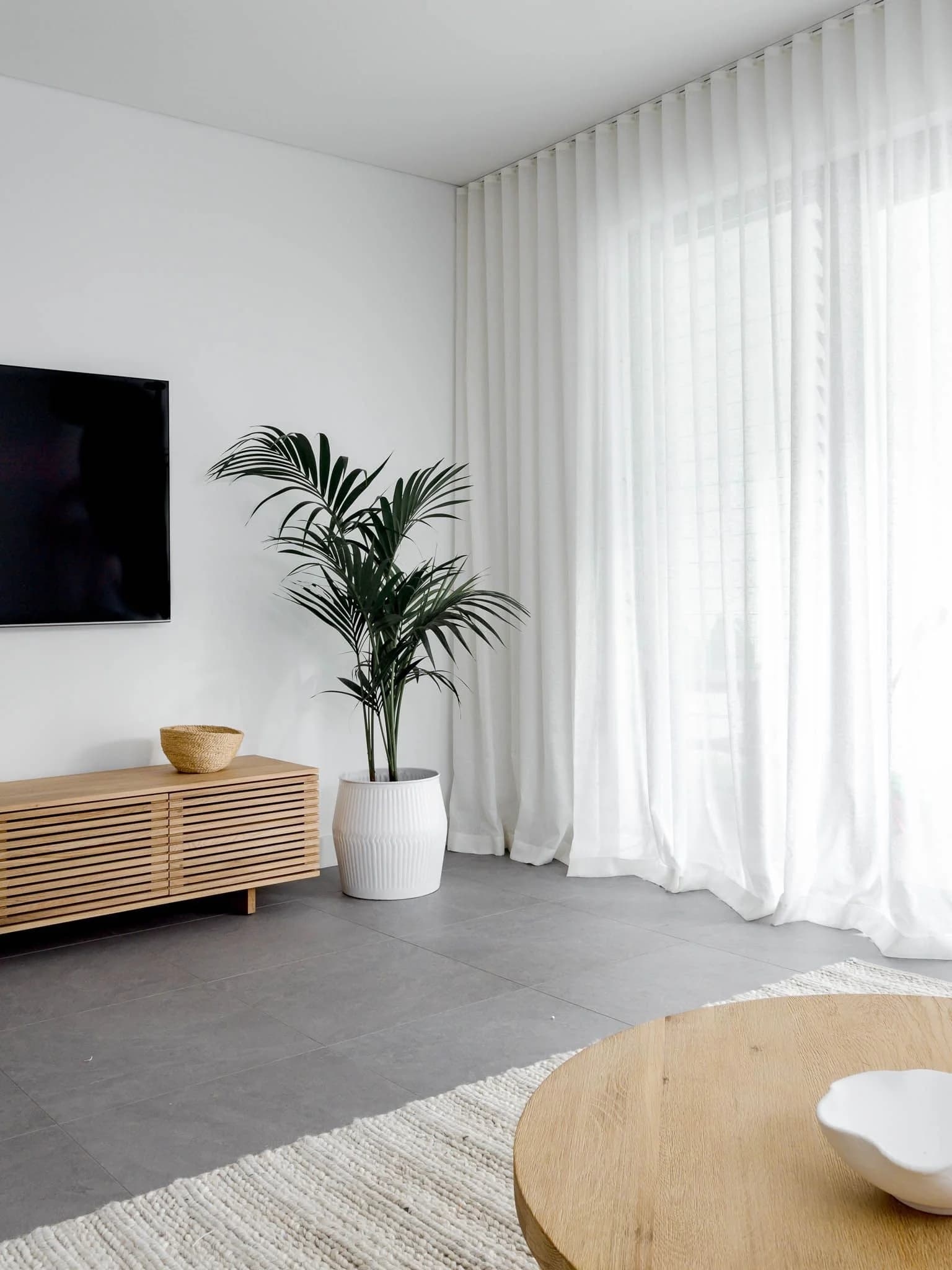 Bright white room with full-height white sheer curtains across a wide window, a palm in a white pot, a slatted wood media cabinet and a round wooden table