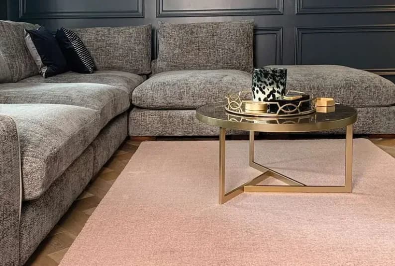Grey textured corner sofa with dark cushions on a plain blush pink rug, a round brass coffee table and dark panelled walls behind