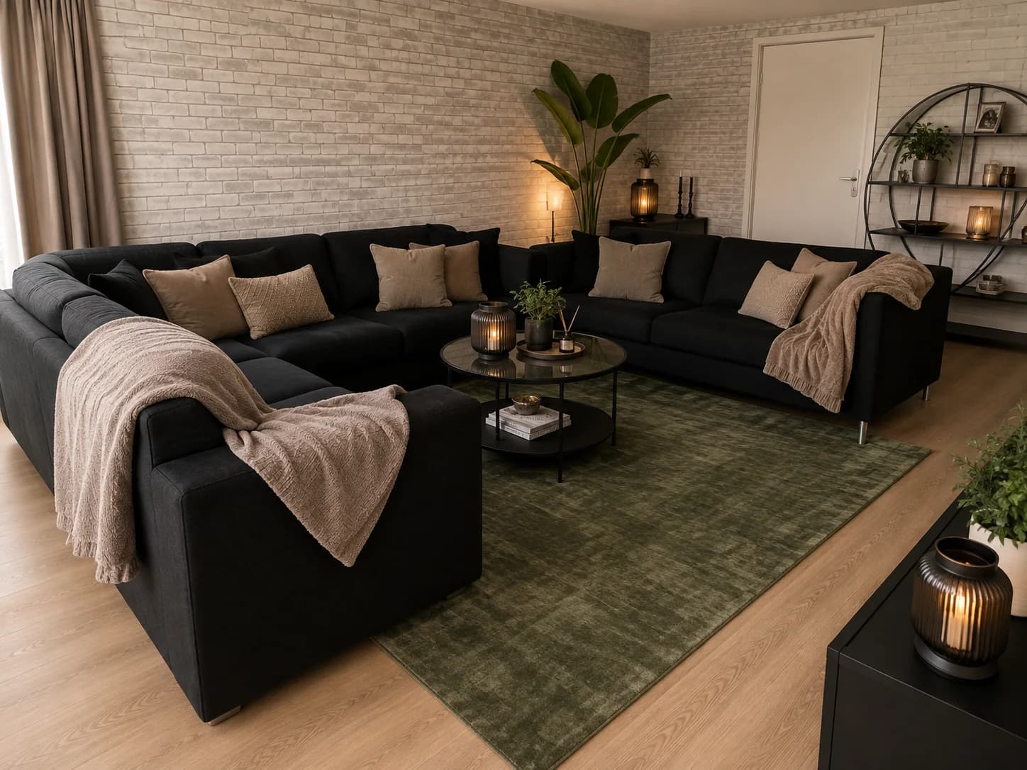 Large black corner sofa with beige cushions and throws on a dark olive green rug over a pale wood floor, with a round glass coffee table and a pale brick wall