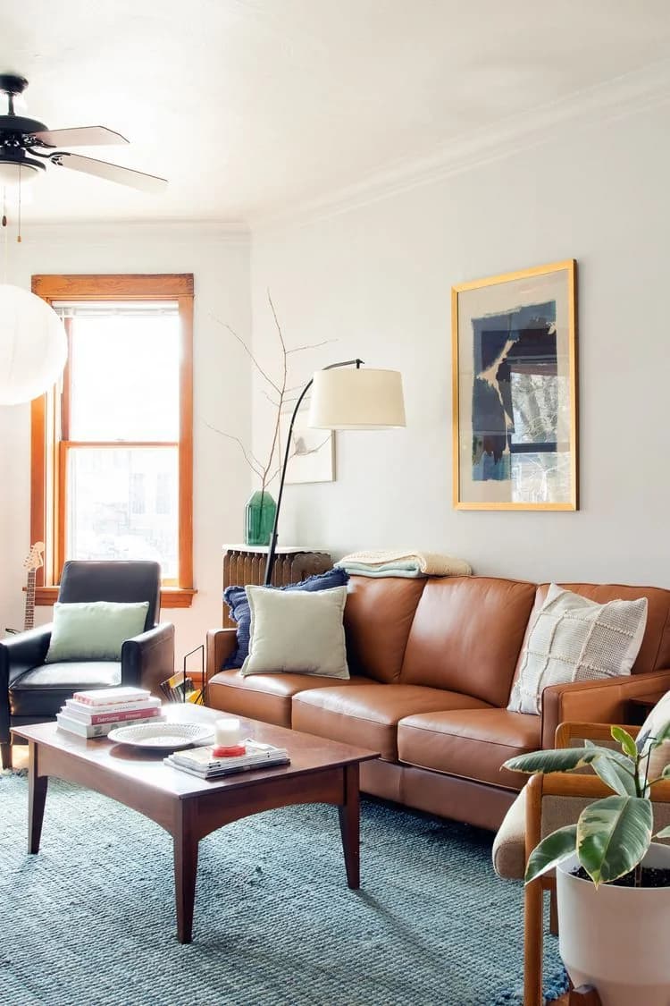 Tan leather sofa with pale cushions on a textured teal blue rug, a dark wood coffee table, a white floor lamp and a framed blue print