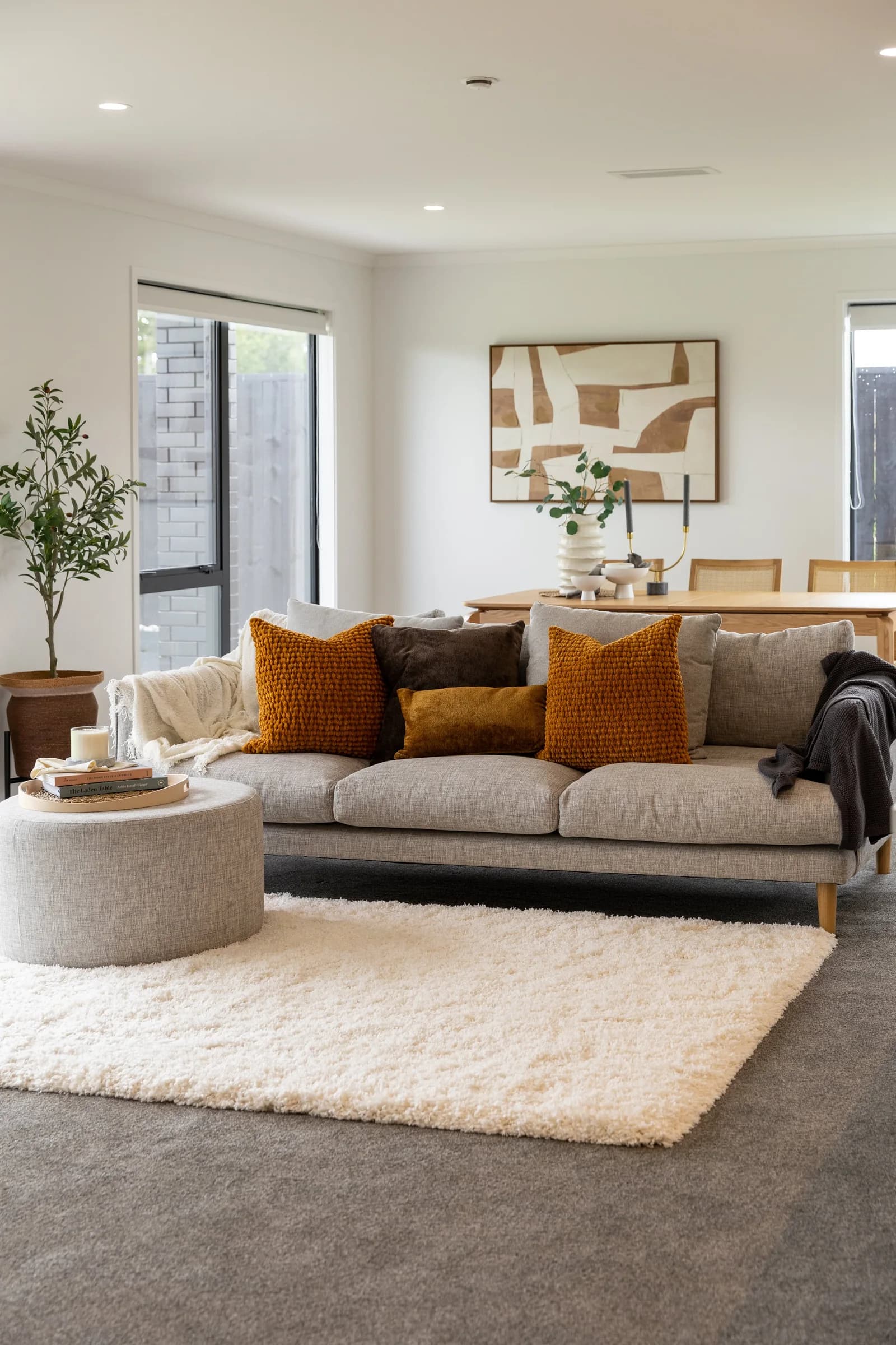 Grey sofa with orange, brown and mustard cushions on a fluffy cream shag rug laid over a grey floor, with a round boucle pouf and a wooden dining area behind