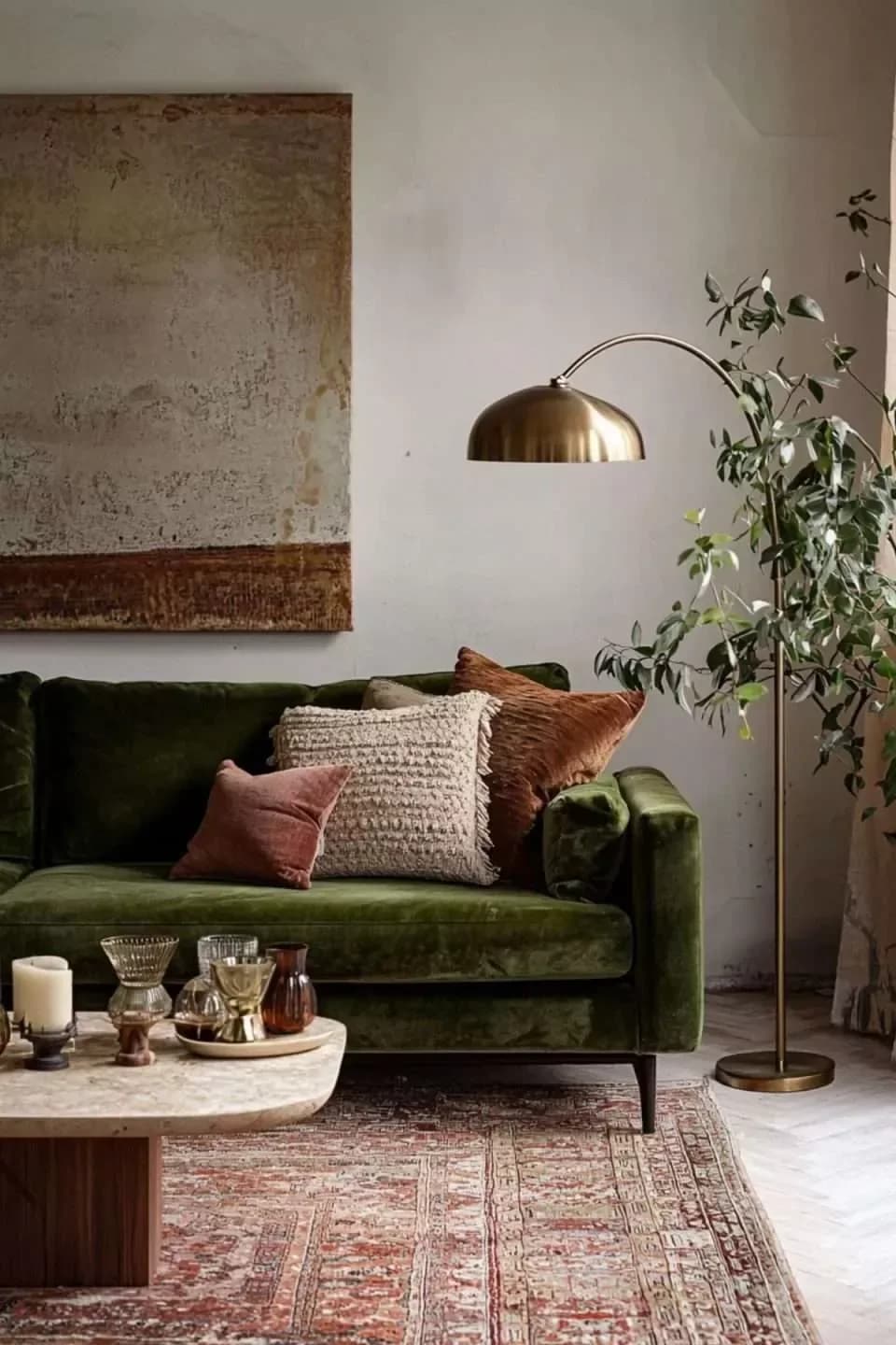 Olive green velvet sofa with rust and cream cushions on a faded red Persian-style rug, with a brass arc floor lamp, a stone coffee table and an olive tree