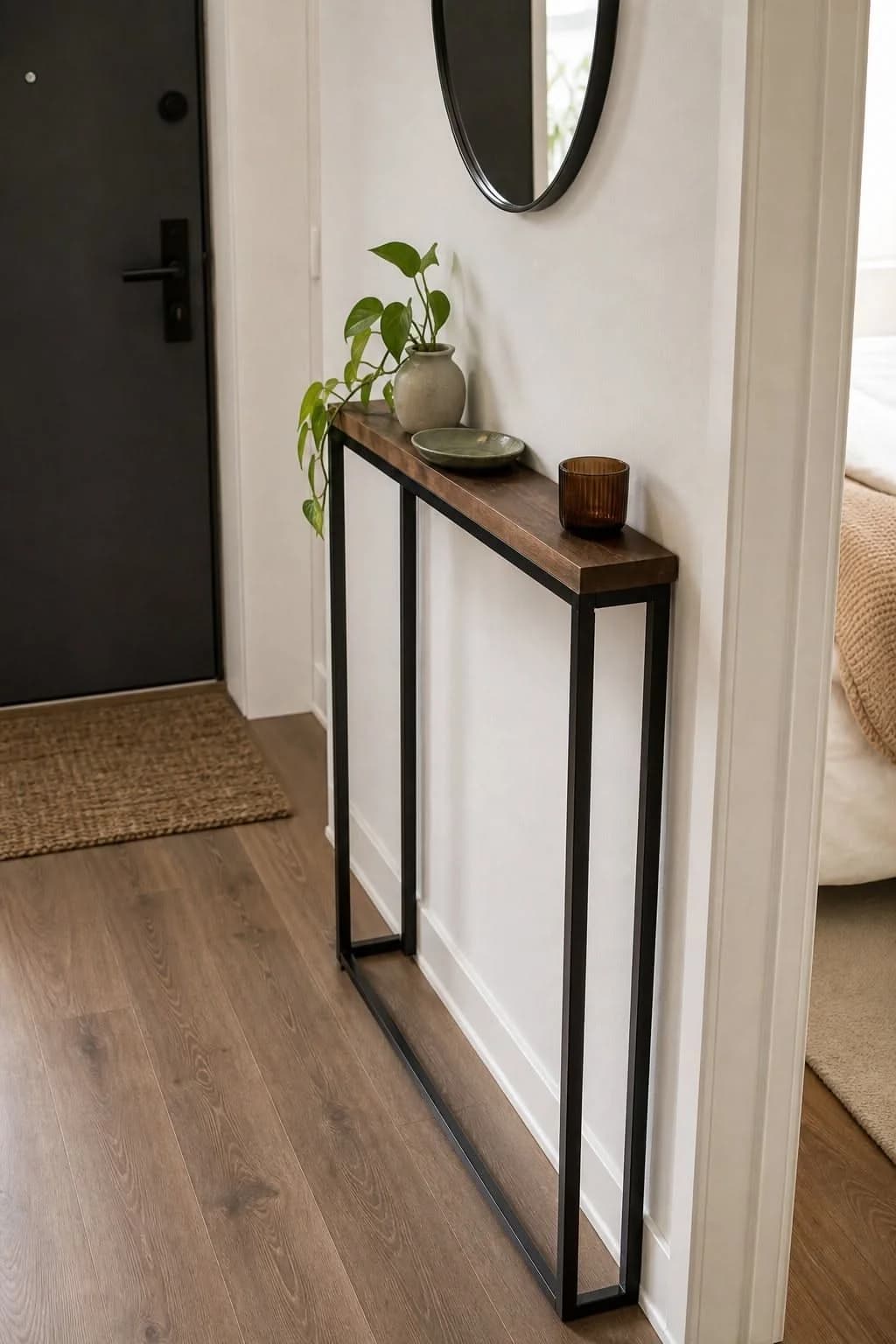 Very narrow black metal console with a thin wooden top beside an apartment front door, holding a small trailing plant, a dish and a candle, with a round mirror above